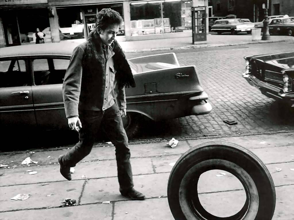 Bob Dylan preparing to kick a tire that is rolling down the sidewalk.