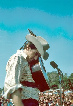Phil Ochs performing live standing by a microphone with his acoustic guitar held to his face.